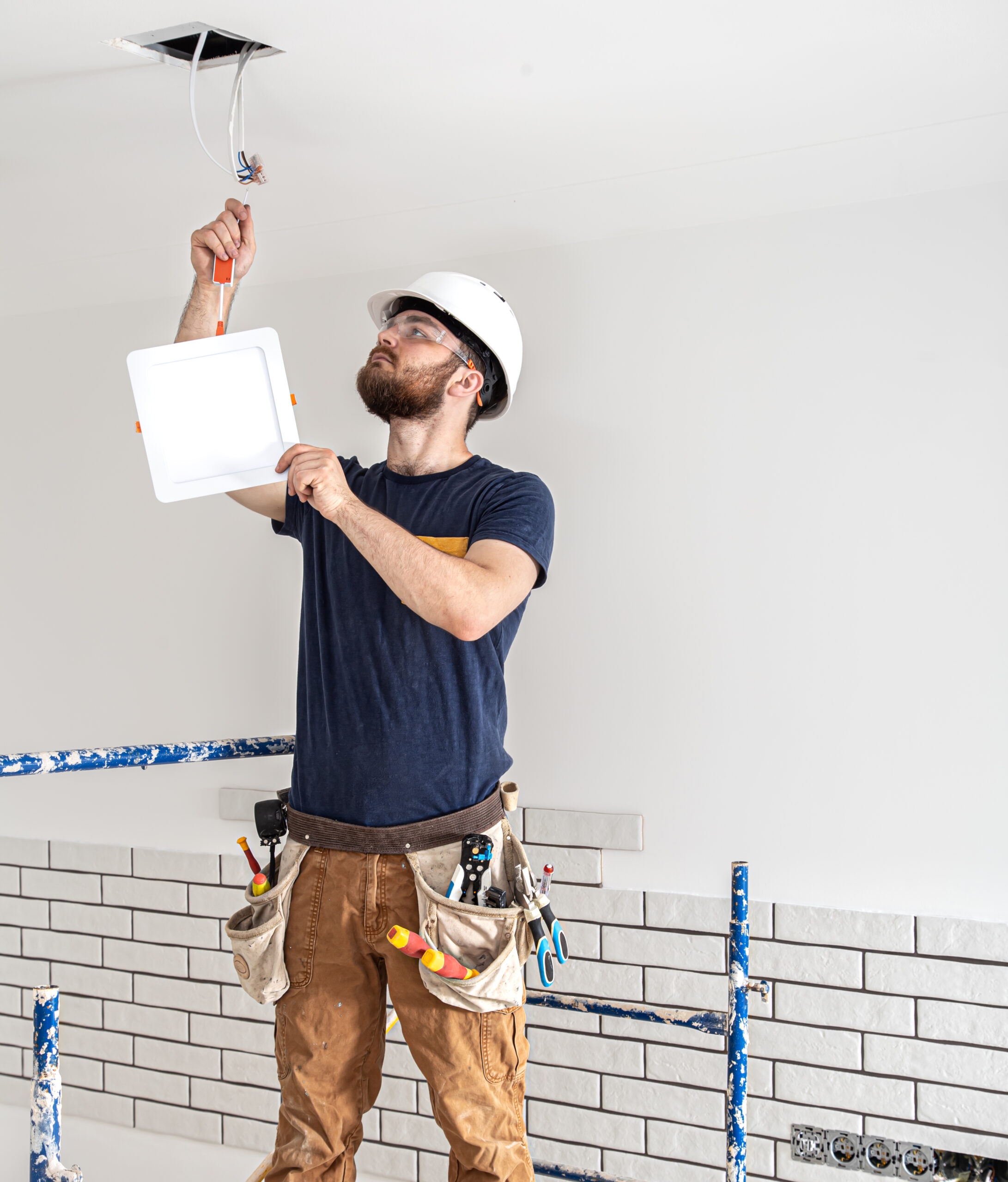 electrician builder with beard worker white helmet work installation lamps height professional overalls with drill background repair site 1 scaled